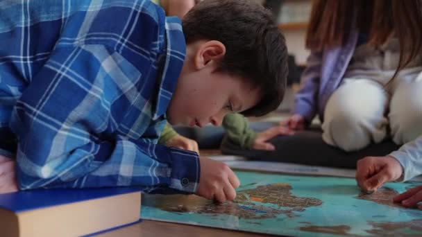 集中した子供は図書館の床に座っているコインと世界地図上のポイントをマークします — ストック動画