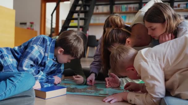 集中注意力的孩子和他们的老师在世界地图上用硬币在图书馆的地板上标出分数 — 图库视频影像