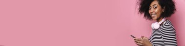 Pembe arka planda izole edilmiş cep telefonu kullanan kulaklıklı neşeli Afrikalı Amerikalı kız.