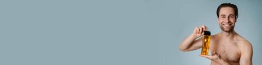 Mavi arka planda izole edilmiş şampuan şişesini sunan gülümseyen genç ve çıplak bir adamın yakın çekimi.