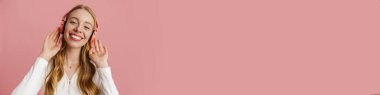Pembe arka planda izole edilmiş kablosuz kulaklıklı neşeli sarışın kadın müzik dinliyor.