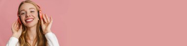 Pembe arka planda izole edilmiş kablosuz kulaklıklı neşeli sarışın kadın müzik dinliyor.