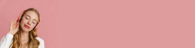 Pembe arka planda izole edilmiş kablosuz kulaklıklı neşeli sarışın kadın müzik dinliyor.