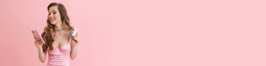 Pembe elbiseli, uzun sarı saçlı, gülümseyen beyaz kadın Barbie iki kredi kartı gösteriyor ve elinde cep telefonu var.