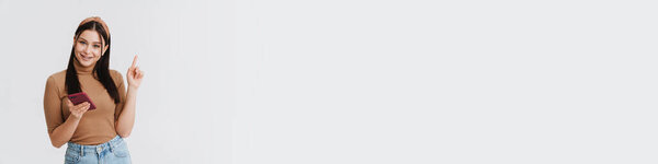 Портрет улыбающейся юной белой брюнетки случайная женщина, стоящая на белом фоне стены держа мобильный телефон указывая в сторону на копировальное пространство