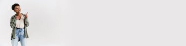 Happy casually dressed young african woman standing over white wall background, looking aside at copy space
