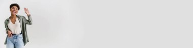 Happy casually dressed young african woman standing over white wall background, looking aside at copy space, waving