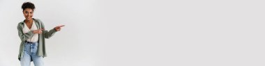 Happy casually dressed young african woman standing over white wall background, pointing aside at copy space