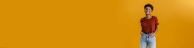 Happy casually dressed young african woman standing over yellow wall background, looking at camera