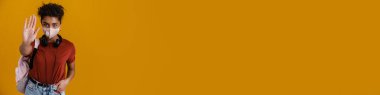 Young black woman in face mask showing stop gesture isolated over yellow background