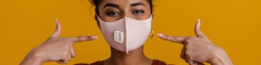 Black woman looking at camera while pointing finger at her face mask isolated over yellow background