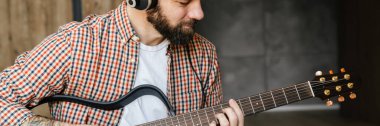 Orta yaşlı, esmer, sakallı bir adam oturma odasında yerde otururken akustik gitar çalıyor ve kulaklık takıyor.