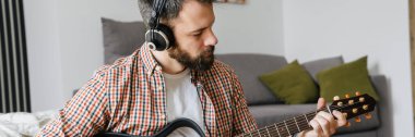 Orta yaşlı, esmer, sakallı bir adam oturma odasında yerde otururken akustik gitar çalıyor ve kulaklık takıyor.