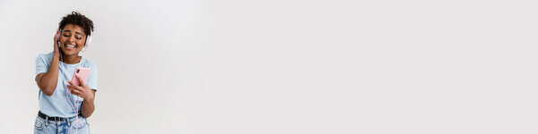 Смеющаяся черная женщина слушает музыку с наушниками и мобильным телефоном изолированы на белом фоне