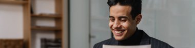 Orta yaşlı, esmer, takım elbiseli bir girişimci ofiste çalışıyor, masada dizüstü bilgisayar ve belgelerle oturuyor, evrak işlerine bakıyor.