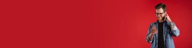 Kırmızı duvar arkasında dikilen, elinde cep telefonu tutan, kablosuz kulaklıkla müzik dinleyen, sıradan giyinen mutlu genç adam.