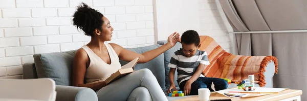 黒女性読書本ながら彼女の息子遊びとともにおもちゃで家 — ストック写真