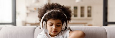 Siyah kıvırcık çocuk evde kanepede otururken kulaklıkla müzik dinliyor.