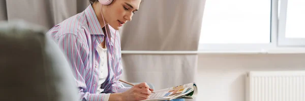 stock image A woman in headphones writing something while sitting with her legs on the sofa in a bright room