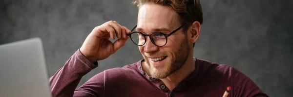 スタジオのテーブルに座っている間にノートパソコンの画面を見て 眼鏡を調整する笑顔の男 — ストック写真