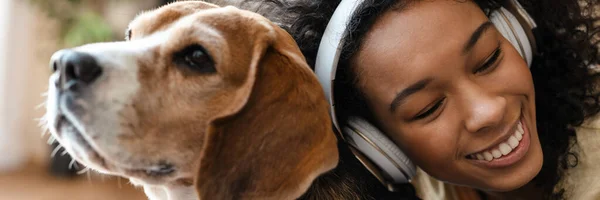 Happy young african woman in headphones relaxing on a couch with her pet dog at home, cuddling