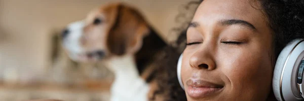 Happy young african woman in headphones relaxing on a couch with her pet dog at home, eyes shut