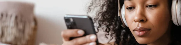 stock image Smiling mid aged african woman listening to music with headphones while relaxing on bed at home, looking at mobile phone in her hands