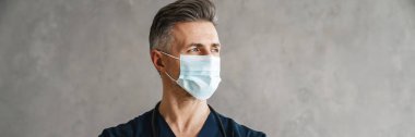 Medikal koruyucu maske takan orta yaşlı bir doktor. Kameraya bakıyor. Kapalı yerde, kolları bağlı.