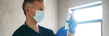 Koruyucu maske takan orta yaşlı erkek doktor elinde aşıyla bir şırınga tutuyor.