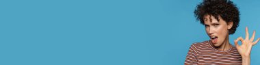 Genç, kıvırcık, heyecanlı kadın, mavi arka planda izole edilmiş kameraya işaret veriyor.