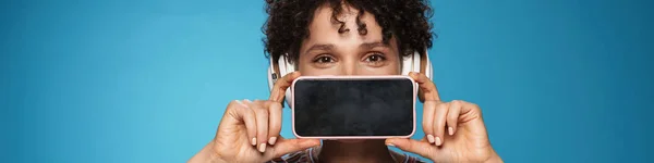 年轻的卷曲黑发女人戴着耳机 显示孤立的蓝色白色背景手机 — 图库照片