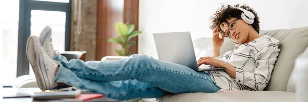 stock image Young curly woman in headphones using laptop while sitting on couch at home