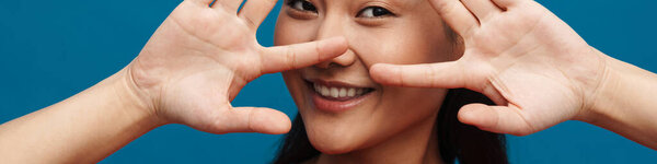 Happy young asian woman in casual clothes standing over blue wall background, covers face with hands