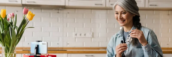 stock image Smiling grey woman showing costume jewelry while taking selfie footage on cellphone at home