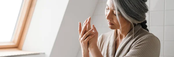 stock image Mature grey woman applying cream while sitting in bathroom at home