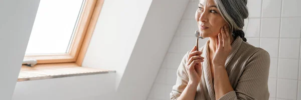 stock image Mature grey woman smiling while massaging face with jade roller in bathroom at home