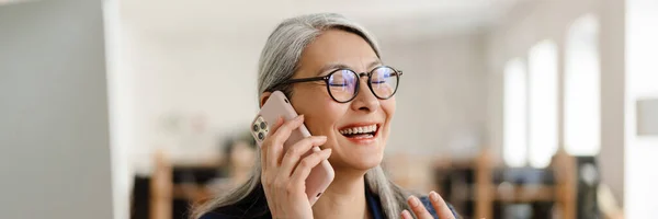 stock image The happy Asian woman in glasses and suit talking on the phone and laughing in a light office