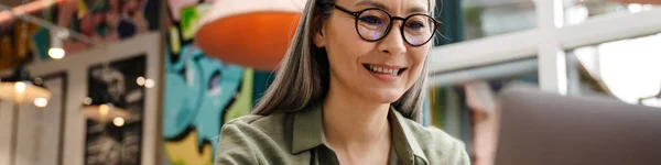 Kypsä Harmaa Nainen Hymyilee Työskentelee Kannettavan Tietokoneen Kanssa Istuessaan Kahvilassa — kuvapankkivalokuva