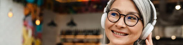Dojrzałe Uśmiechnięta Kobieta Słuchanie Muzyki Słuchawkami Komórką Kawiarni Wewnątrz — Zdjęcie stockowe