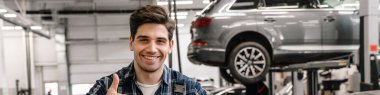 Genç araba tamircisi garajda dikilirken gülümsüyor ve başparmağını gösteriyor.