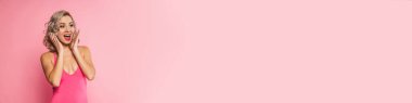 Yüzünde mayo olan gülümseyen sarışın bir kadının portresi pembe duvarın arkasında duruyor, yan bakıyor.