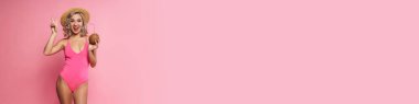 Mayo giymiş, elinde hindistan cevizi kokteyli tutan, pembe arka planda yalnız poz veren mutlu sarışın kadın.