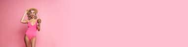 Mayo giymiş, elinde hindistan cevizi kokteyli tutan, pembe arka planda yalnız poz veren mutlu sarışın kadın.