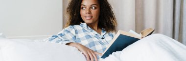 Yatakta uzanmış kitap okuyan gülümseyen Afrikalı bir kadın.