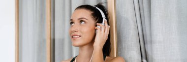 Spor kıyafetli, kulaklıklı, müzik dinleyen, oturma odasında cep telefonu tutan gülümseyen beyaz bir kadın.