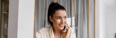 Beyaz esmer kadın evde ödev yaparken cep telefonuyla konuşuyor.