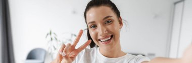 Gülümseyen esmer kadın evde selfie çekerken barış işareti gösteriyor.