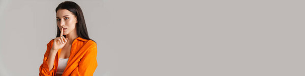 Молодая брюнетка в рубашке с жестом молчания на сером фоне