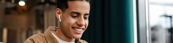 Молодой Улыбчивый Латиноамериканец Сидит Столом Кафе Ноутбуком Помещении Держит Мобильный — стоковое фото