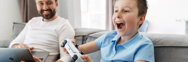 自宅でビデオゲームをプレイするゲームパッドを持つ幸せな父親と小さな息子 ラップトップコンピュータで作業している父 — ストック写真